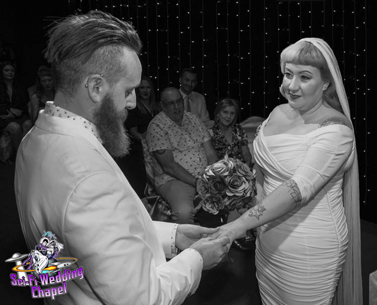 Black and white image of groom putting wedding ring on bride with attendees in the background at Sci-Fi Wedding Chapel.